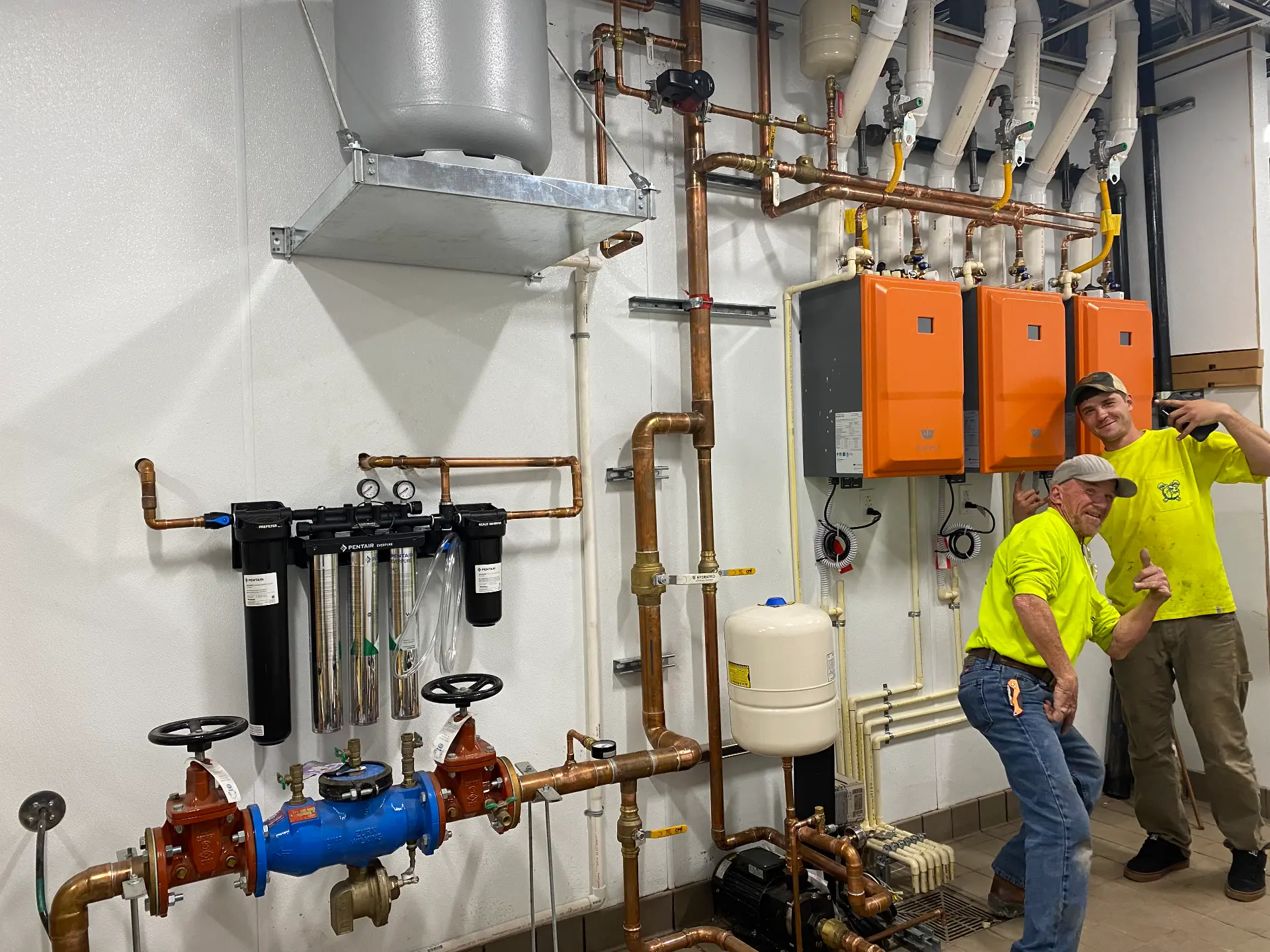Littles Plumbing technicians beside a finished mechanical room with copper manifolds, tankless water heaters, filtration, and a backflow preventer